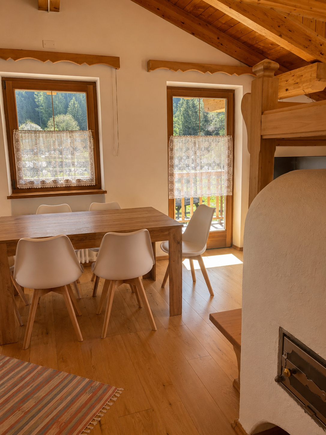 Zona pranzo luminosa con travi a vista, finestre panoramiche e accesso al balcone est con vista Dolomiti